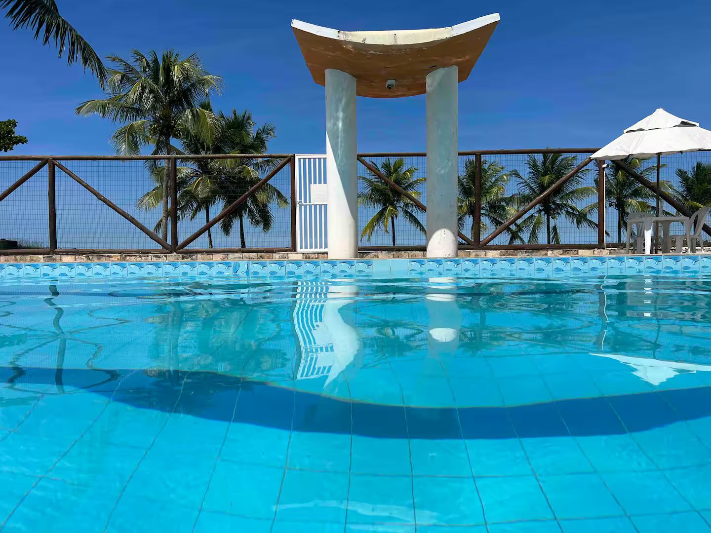 A clear blue swimming pool with a white structure in the center, surrounded by a fence and palm trees.