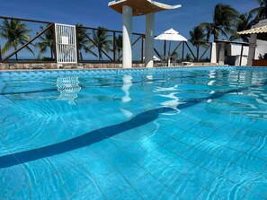 En klar, blå pool med flisekant, omgivet af et hegn, palmer og en hvid pavillon.