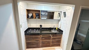 A compact kitchen with a black countertop, a sink, and a built-in stove. There is a wooden shelf above the countertop and a mirror on the wall. - Apartment on the 1st floor facing the sea on Guaibim beach. (Guaibim)