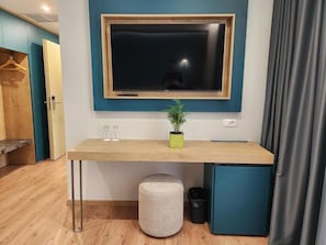 Una habitación con un escritorio de madera, un televisor montado en la pared, una planta en maceta y un cesto de basura.
