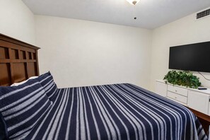 Ein Schlafzimmer mit einem Bett, einem Holzkopfteil, einer weißen Kommode, einem Flachbildfernseher und einer Topfpflanze.