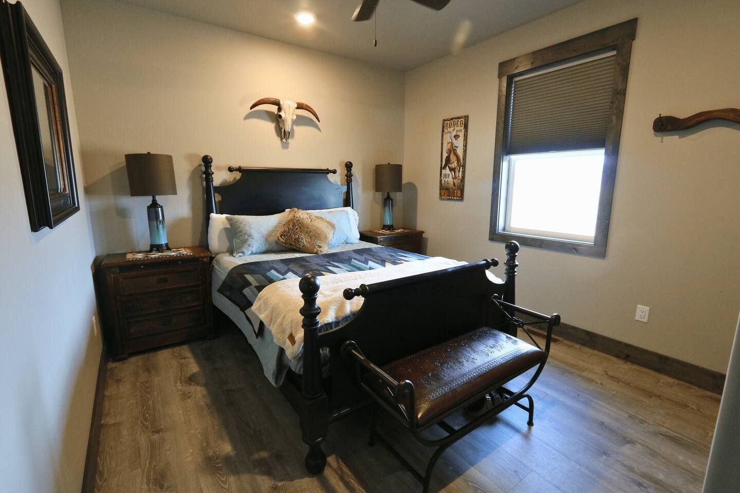 Une chambre à coucher comprenant un lit, un banc, une table de chevet, une lampe, une décoration en forme de crâne fixée au mur, et une fenêtre avec des stores.