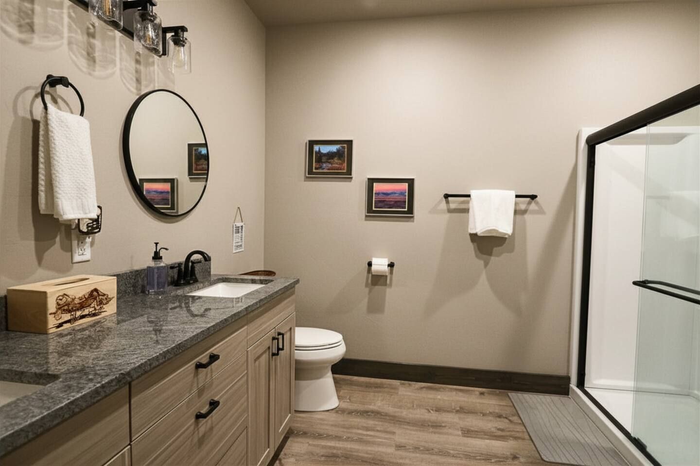 A modern bathroom with a granite countertop, a white toilet, a glass shower enclosure, and framed pictures on the wall.