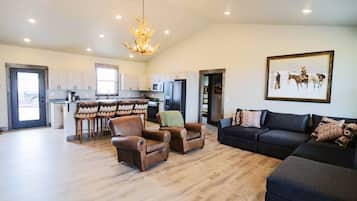 Een ruime woonkamer met een donkergrijze bank, twee leren fauteuils, een kroonluchter en een eethoek met een barkrukset.