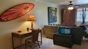 A living room with a desk, a chair, a sofa, a lamp, a colorful painting, and a ceiling fan. - Exceptional Family 2-bed/2 bath AC Pool & Garden View on Oceanfront Property (Lahaina)