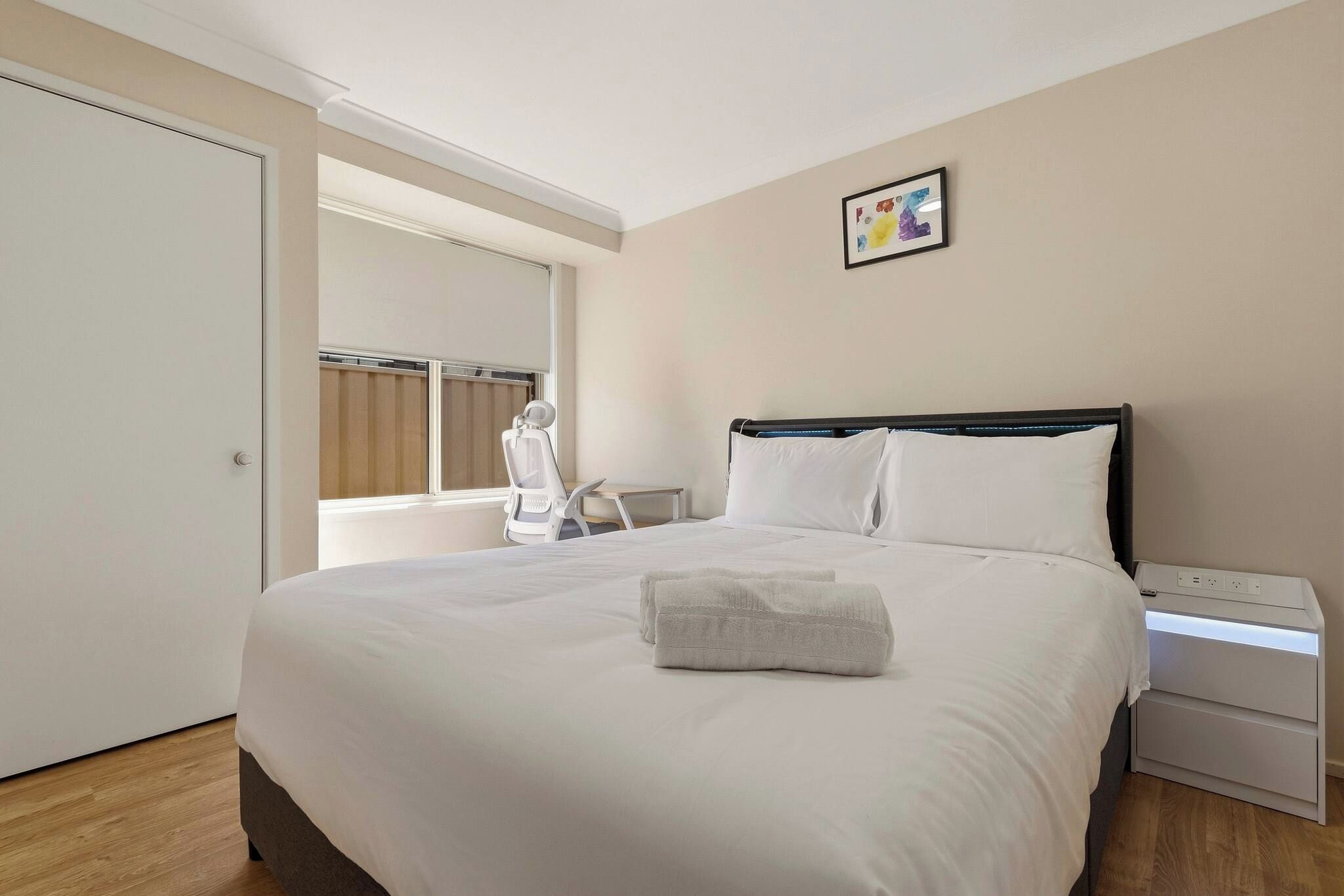 A hotel room with a bed, a bedside table, a nightstand, a window with blinds, and a door.