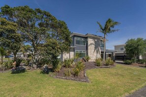 A modern two-story house with a landscaped front yard featuring a variety of plants and trees. - Peaceful Paradise | The Family Retreat (Whitianga)