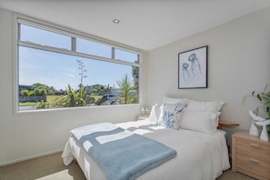A bedroom with a large window, a bed with white and blue bedding, a nightstand, a wooden dresser, and a framed picture on the wall. - Peaceful Paradise | The Family Retreat (Whitianga)