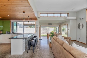 A modern kitchen with a central island, bar stools, and a dining area with a stone wall and outdoor view. - Peaceful Paradise | The Family Retreat (Whitianga)