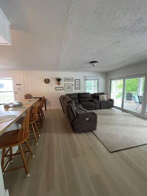Ein modernes Wohnzimmer mit einem Ecksofa, einem Essbereich mit Holzkästen und einem großen Fenster, das für natürliches Licht sorgt.