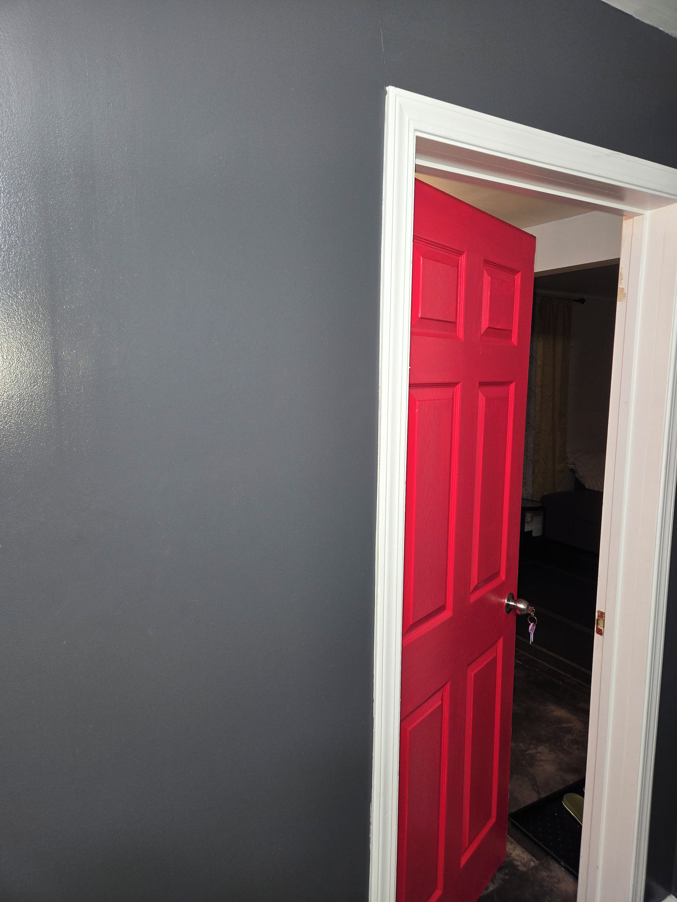 A red door with a white frame, leading to a room with a visible bed and a dark-colored wall.