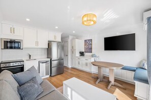 A modern kitchen with white cabinetry, stainless steel appliances, and a dining area with a wooden table and blue upholstered seating. - Heron's Hideaway / Ocean View, Steps to the Beach (Hilton Head Island)