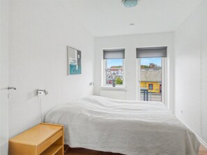 Une chambre à coucher avec un lit, une table de chevet, une fenêtre donnant sur des immeubles et un luminaire au plafond.