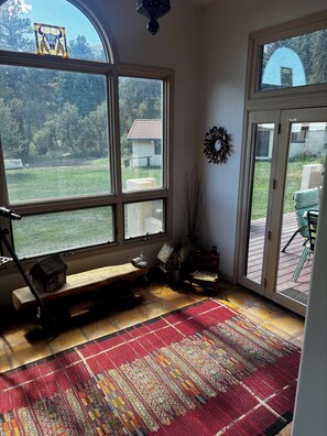 Ein Zimmer mit großen Fenstern, einer Bank, einem Vogelhäuschen, einem Sessel und einem gemusterten Teppich.