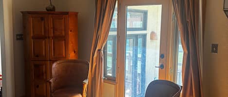 A room with wooden flooring, a wooden cabinet, two chairs, a table, and a view through a door.