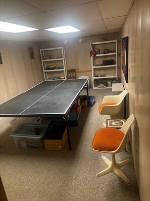 Une pièce comprenant une table de ping-pong, deux chaises, des meubles de rangement et des étagères contenant divers objets.