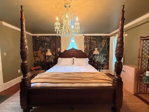 A four-poster bed with a chandelier, a tropical wallpaper, and a nightstand with a lamp.