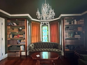 A room with a large, dark-colored sofa, a wooden coffee table, and a chandelier. There are bookshelves on either side of the sofa, and a stained glass window in the center.