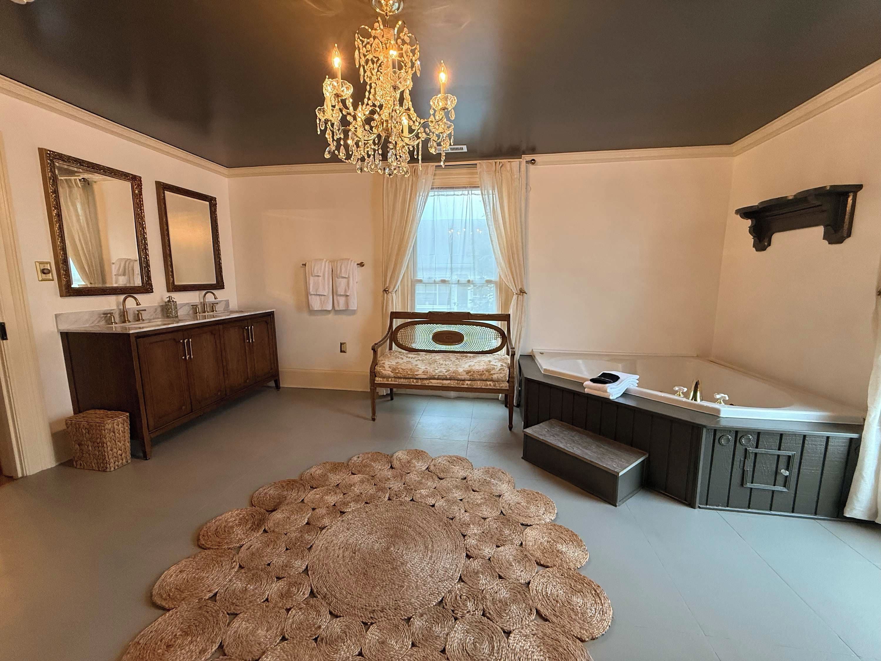 A bathroom with a large bathtub, a vanity with a mirror, a chandelier, and a woven rug on the floor.