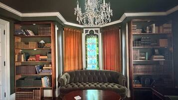 A room with a large, dark-colored sofa, a wooden coffee table, and a chandelier. There are bookshelves on either side of the sofa, and a stained glass window in the center.