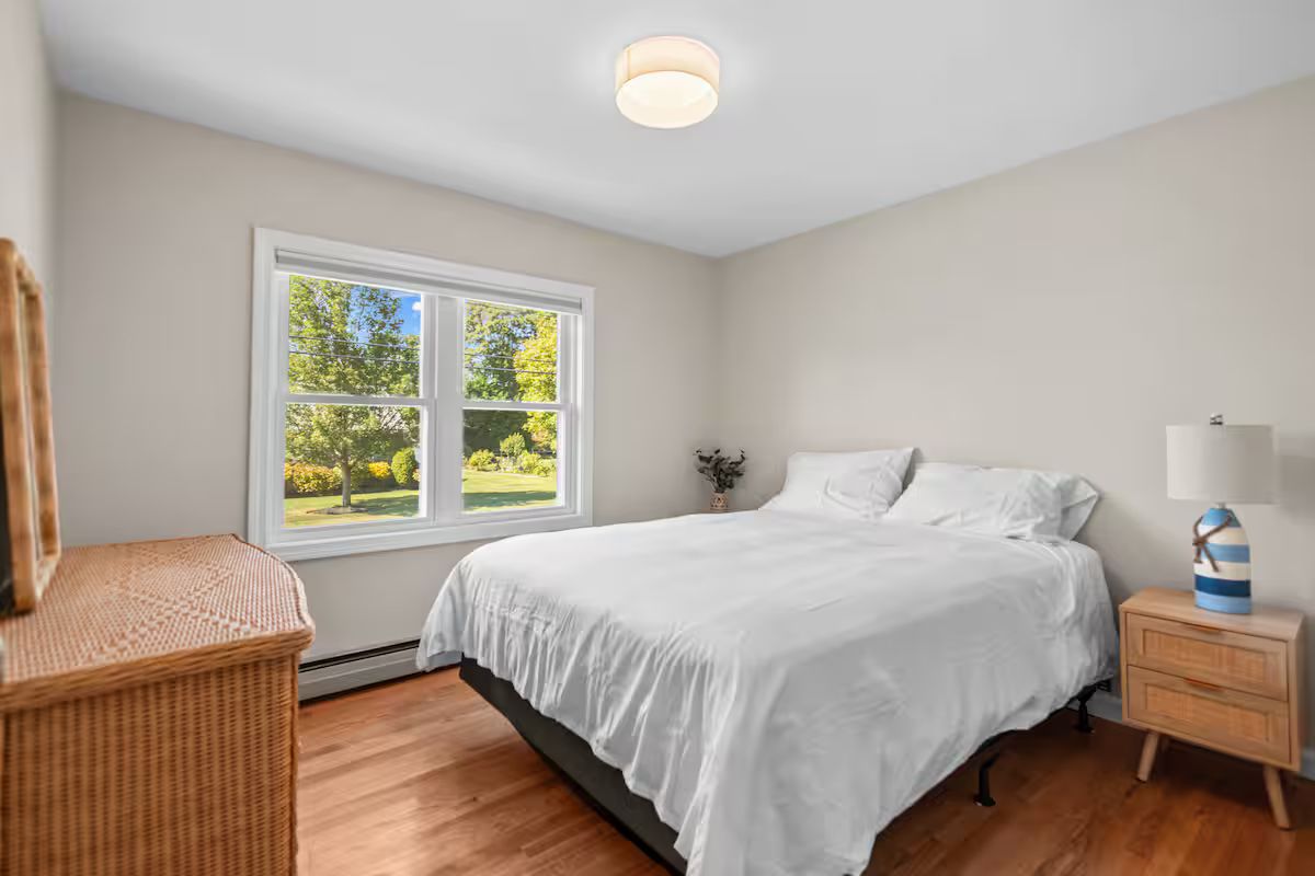 A bedroom with a large window, a bed, a nightstand, and a wicker chair.