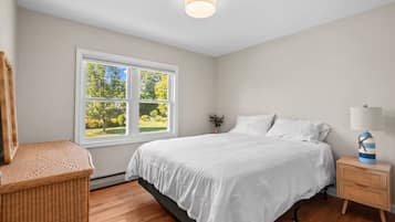 A bedroom with a large window, a bed, a nightstand, and a wicker chair.