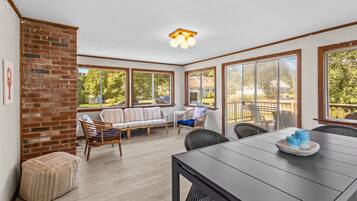 A sunlit room with a brick wall, a sofa, a dining table, and large windows offering a view of the outdoors.