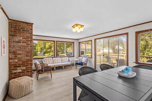 A sunlit room with a brick wall, a sofa, a dining table, and large windows offering a view of the outdoors. - Family Friendly Beach House with linens and AC - Sleeps 8 (Dennis)