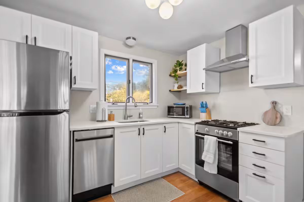 A modern kitchen with white cabinets, stainless steel appliances, and a window with a view of trees.