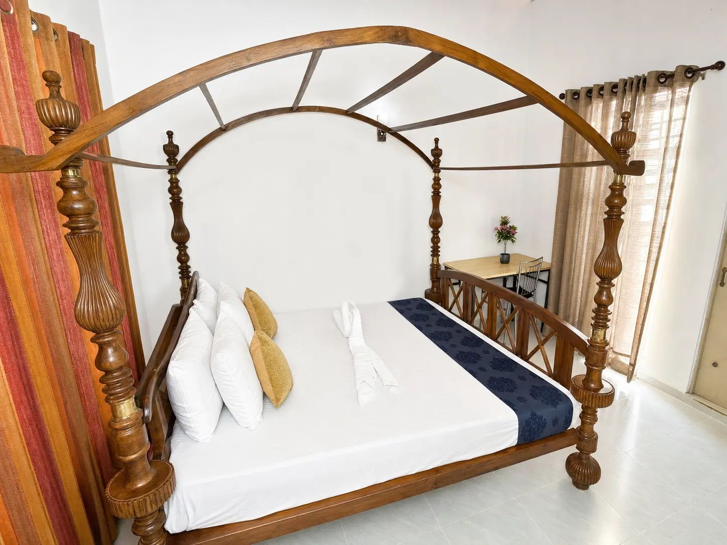 A four-poster bed with a wooden frame and white bedding, a small table with a chair, and a window with curtains.