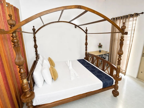 A four-poster bed with a wooden frame and white bedding, a small table with a chair, and a window with curtains.