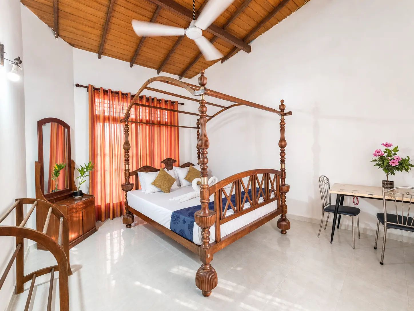 A four-poster bed with a wooden frame, a ceiling fan, and a desk with chairs in a room with white walls and a tiled floor.