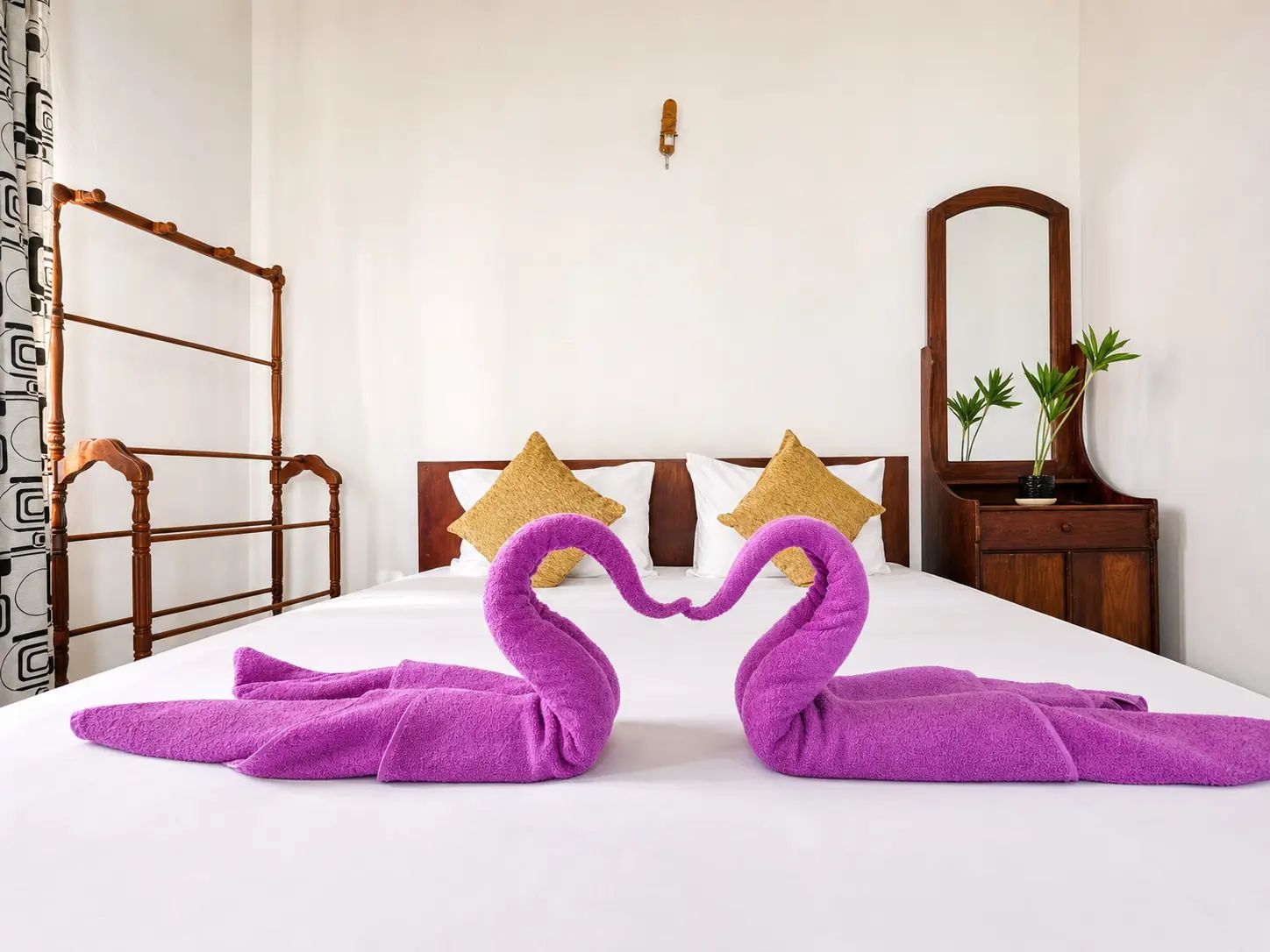 A hotel room with a bed, two pillows, a wooden headboard, a nightstand with a mirror, and a plant.