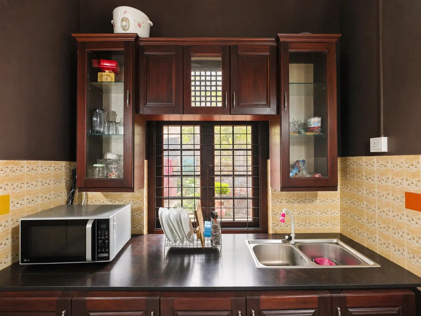 A kitchen with dark wooden cabinets, a microwave, a sink, and a window with a view of greenery.