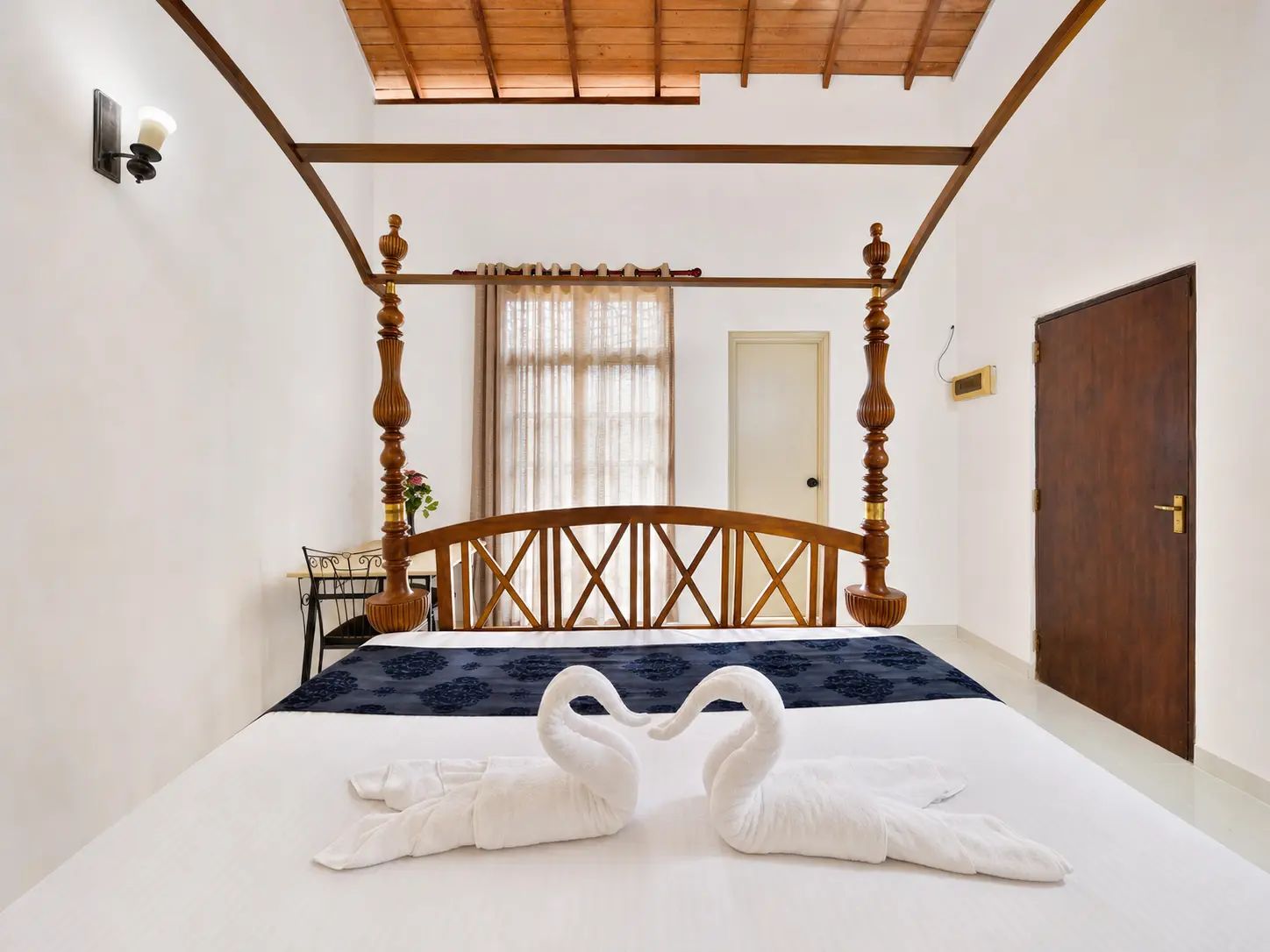 A bedroom with a four-poster bed, a wooden door, and a wall-mounted light fixture.