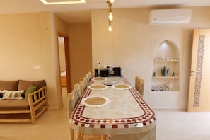 A dining area with a long table, chairs, and a kitchen area in the background. - Authentic Apartment in Tamraght – A Peaceful Stay Near the Beach (Tamraght)