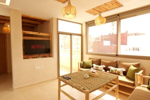 A modern living room with a television mounted on the wall, a wicker table, and a sofa with cushions. - Authentic Apartment in Tamraght – A Peaceful Stay Near the Beach (Tamraght)