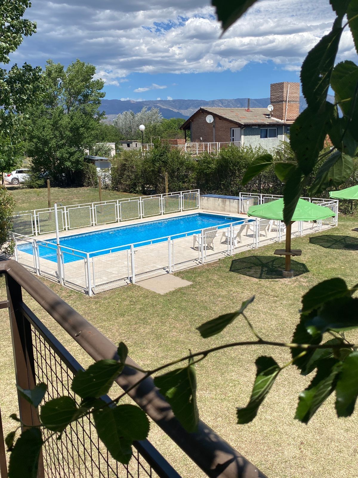 Una piscina rectangular con una valla blanca, rodeada de césped y árboles, con una cadena montañosa al fondo.
