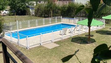 Una piscina rectangular con una valla blanca, rodeada de césped y árboles, con una cadena montañosa al fondo.