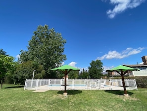 A fenced outdoor area with two green umbrellas, a pool, and a large tree. - Eluney Cabañas (Mina Clavero)