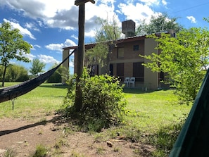A hammock tied to a wooden post in a grassy area with a house in the background. - Eluney Cabañas (Mina Clavero)