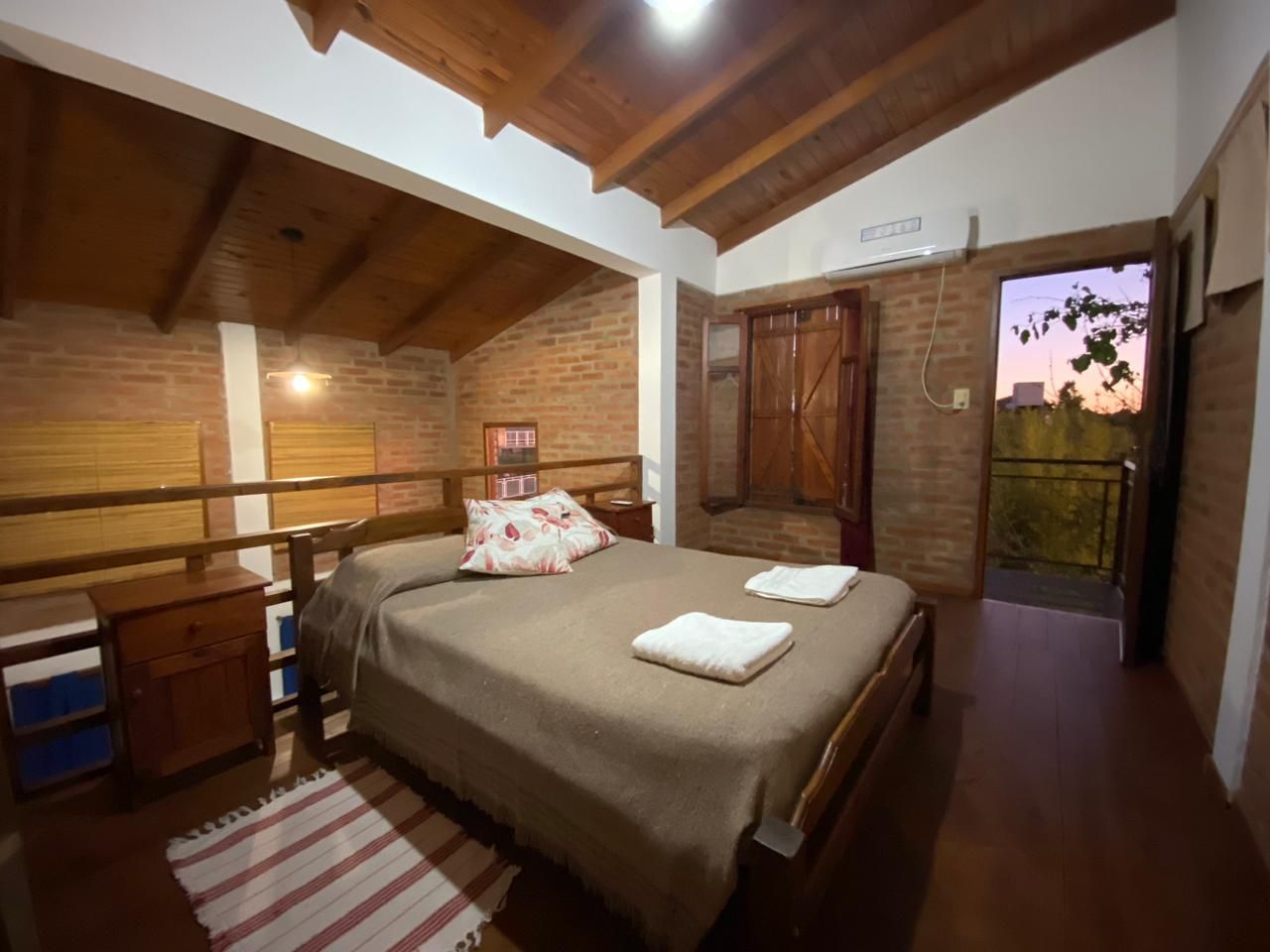 Ein Schlafzimmer mit Holzboden, einem Bett mit brauner Bettdecke, einem Nachttisch, einer Kommode und Blick ins Freie.