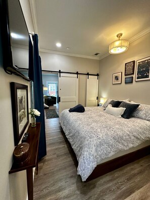 A bedroom with a bed, a bedside table, a lamp, and a door leading to another room. - Quaint and charming 2 bed cottage with a putting green & outdoor shower! (Pensacola)