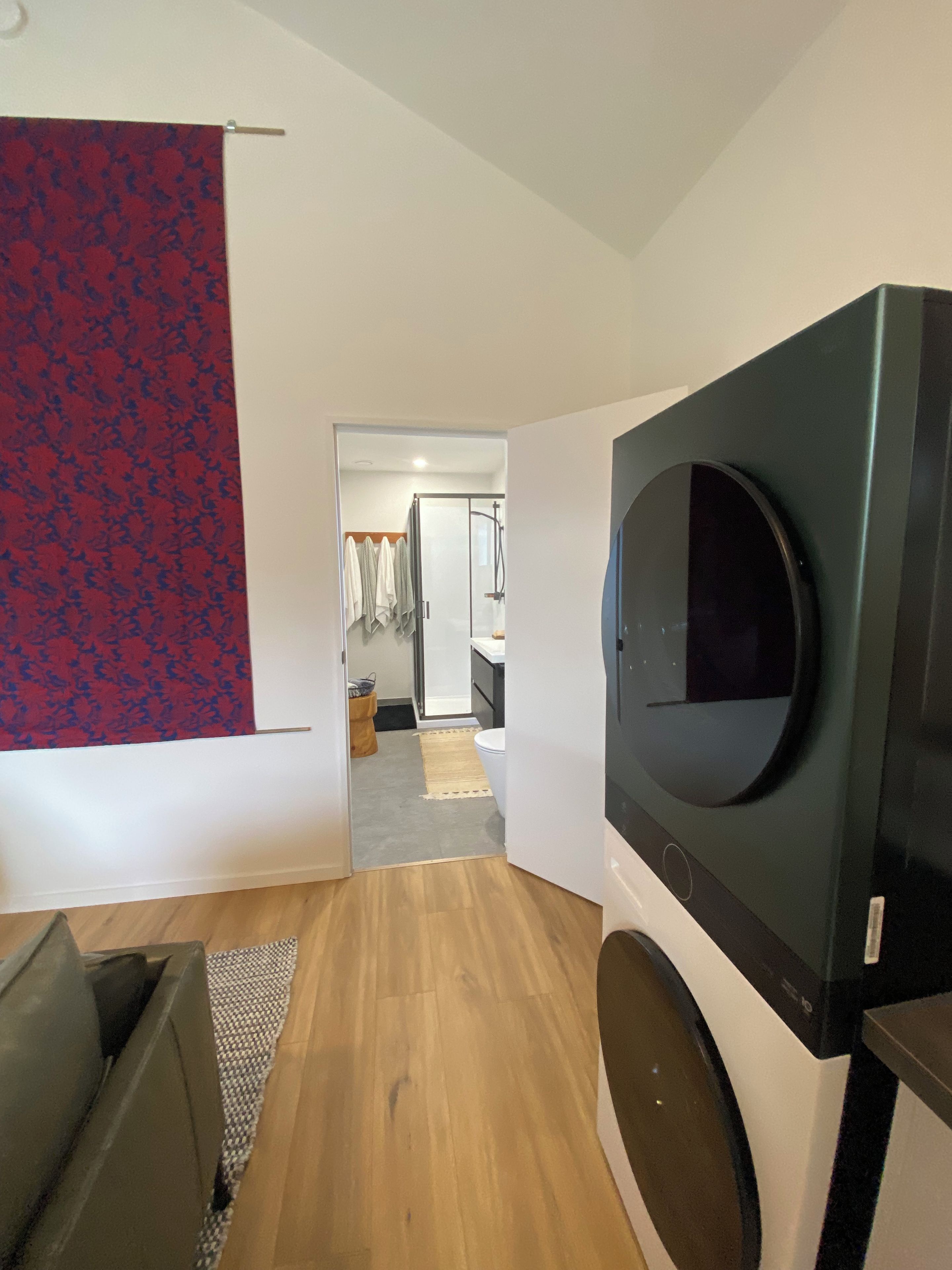 A modern living room with a flat-screen TV mounted on the wall, a wooden floor, and a hallway leading to a bathroom.