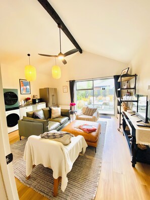 A modern living room with a sofa, a coffee table, and a TV. There is a dining area with a table and chairs. The room has a ceiling fan and a window with curtains.
