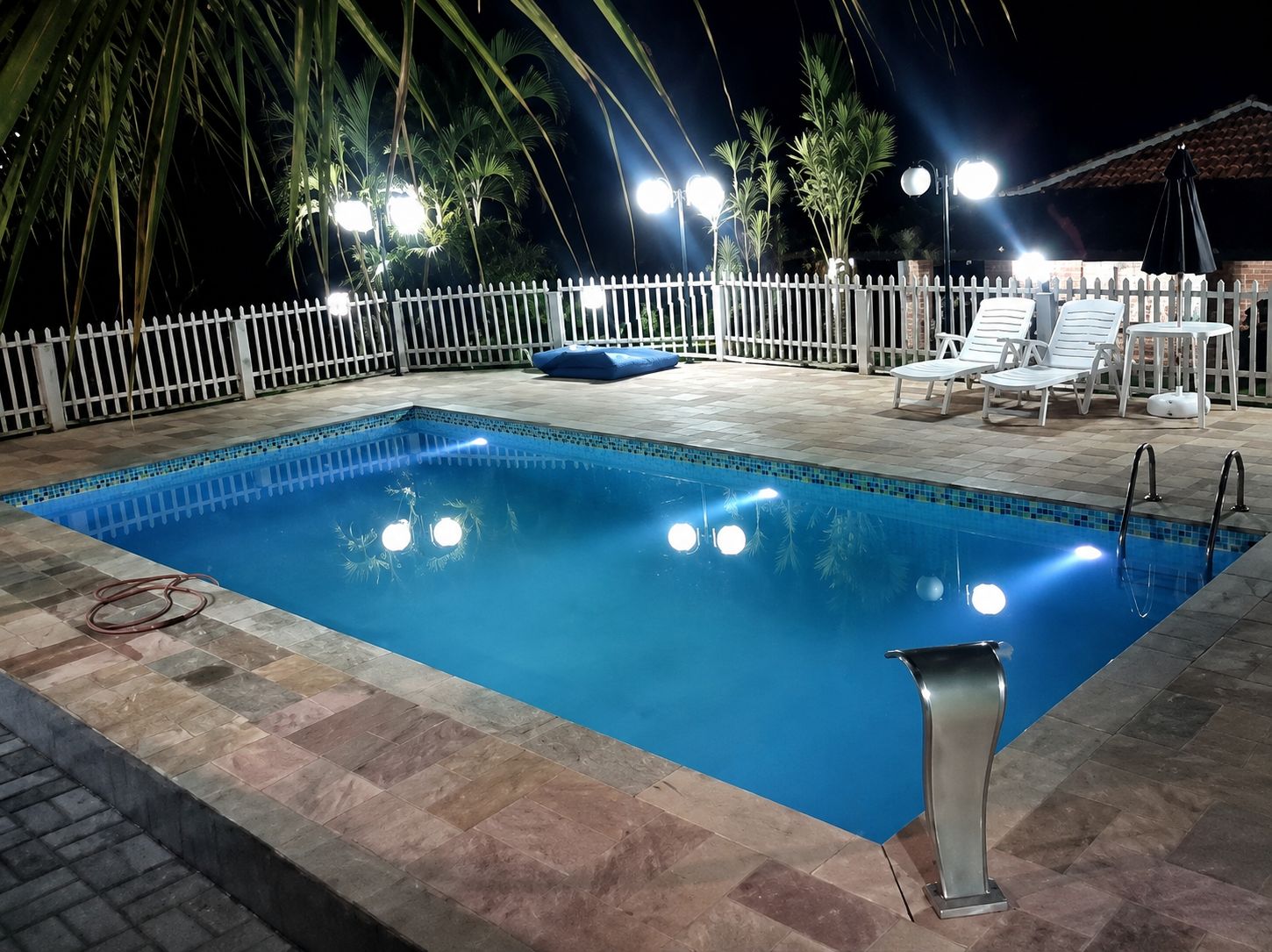 Une piscine rectangulaire dont les bords sont recouverts de carreaux, entourée d’une clôture blanche et équipée d’une échelle en métal.