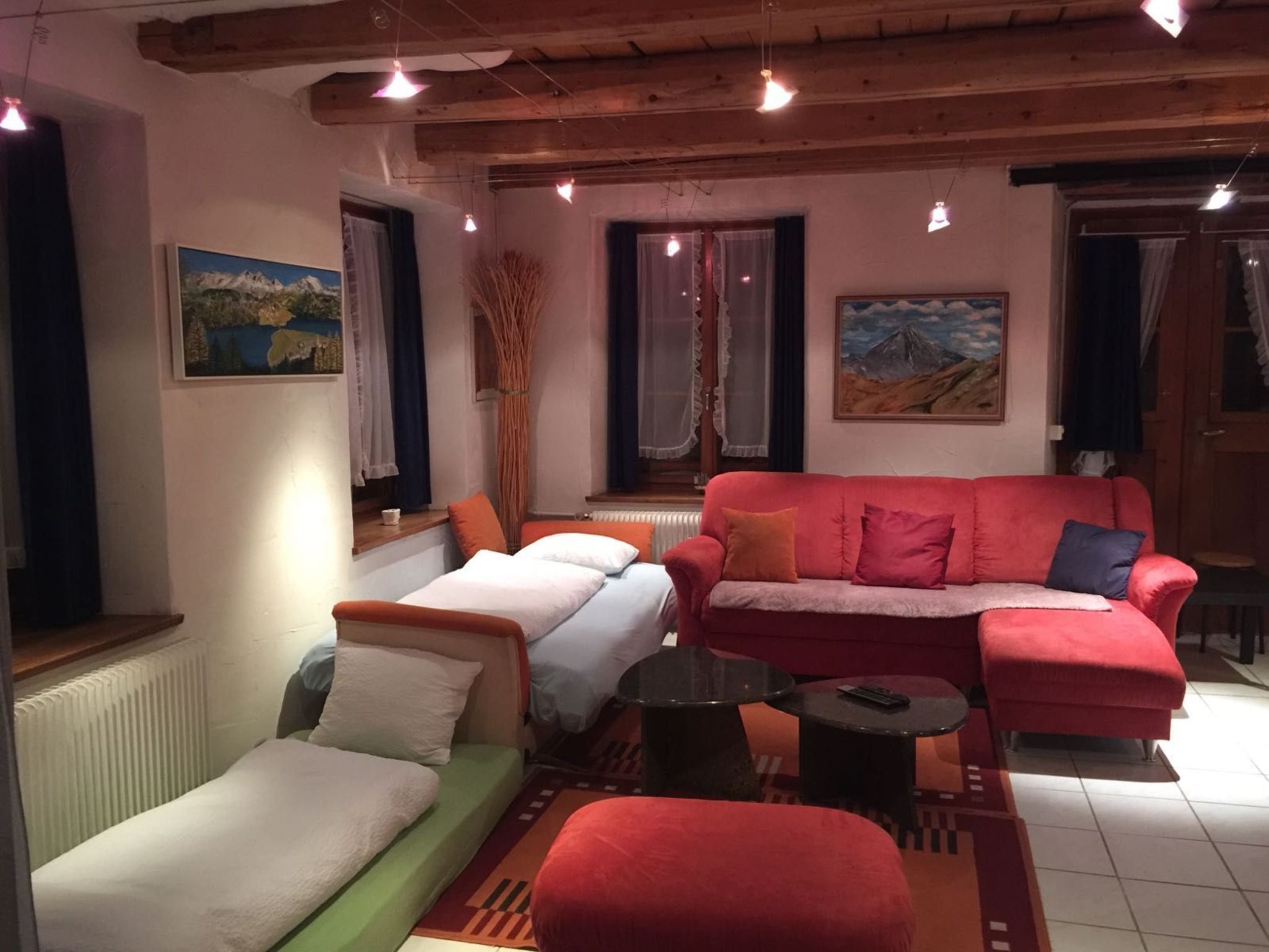 Una stanza accogliente con un divano rosso, un divano verde e un letto piccolo. Sono presenti due finestre con tende, un termosifone e un quadro appeso al muro.
