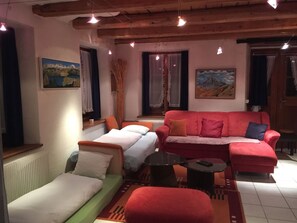 A cozy room with a red sofa, a green sofa, and a small bed. There are two windows with curtains, a radiator, and a painting on the wall. - Haus alte Post, (Samnaun-Compatsch) by Interhome (Samnaun)