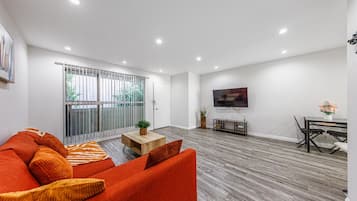 Uma sala de estar moderna com sofá, mesa de centro, televisão na parede e área de jantar com mesa e cadeiras.
