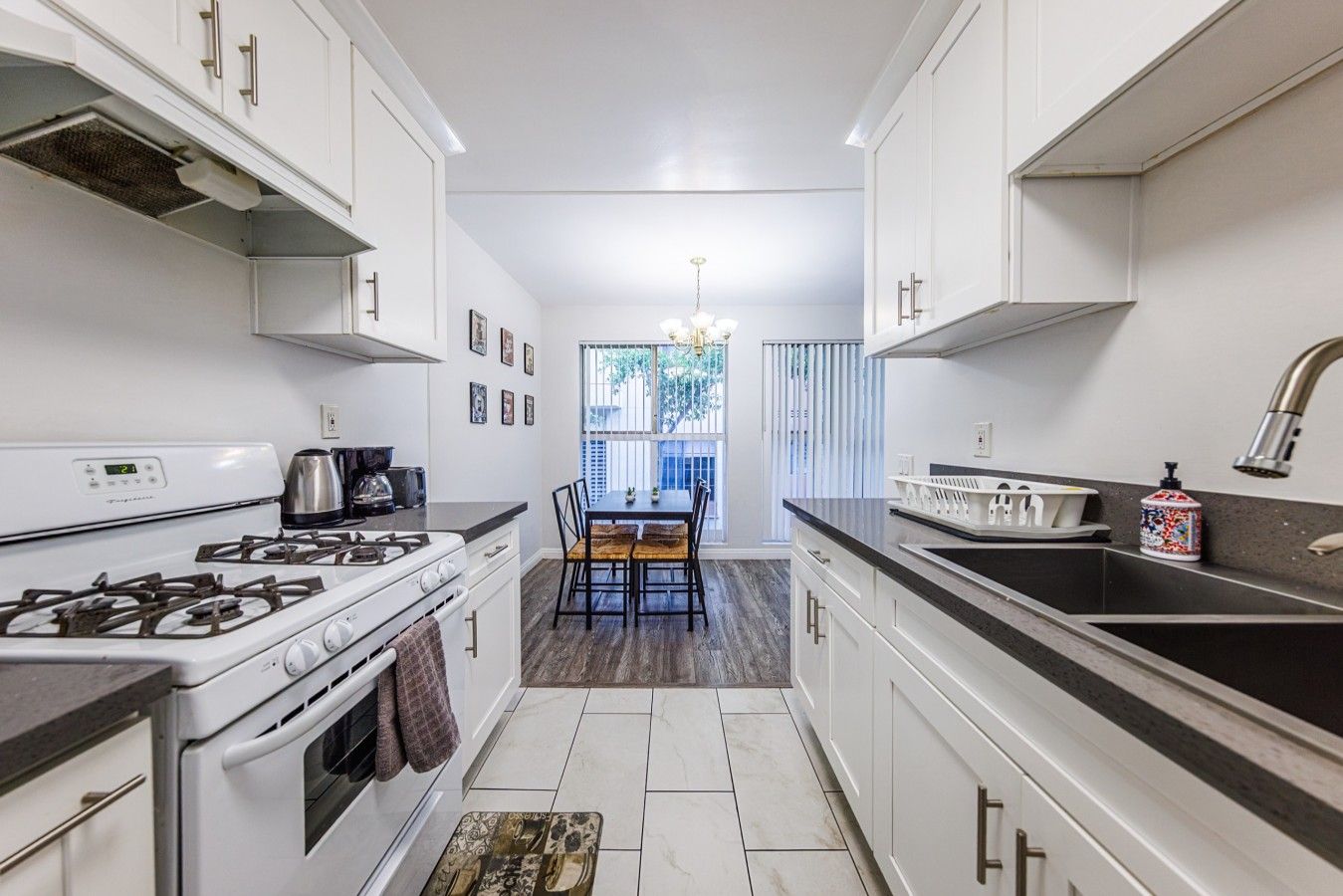A narrow kitchen with white cabinets, a gas stove, a coffee maker, and a dining area with a table and chairs.